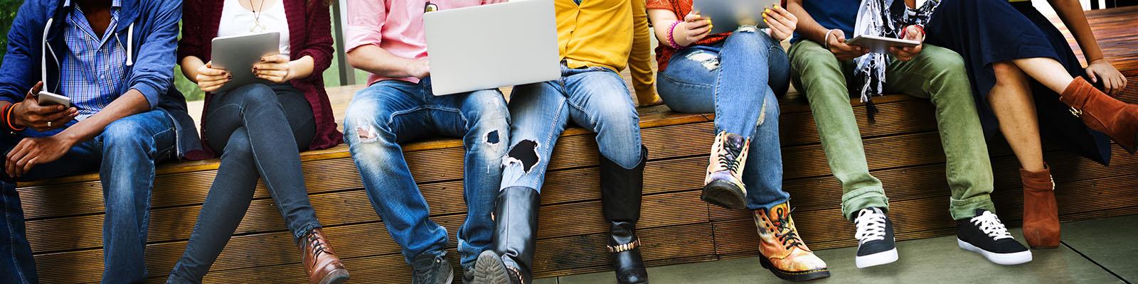 Close up of students sitting on a bench using laptops and other technology